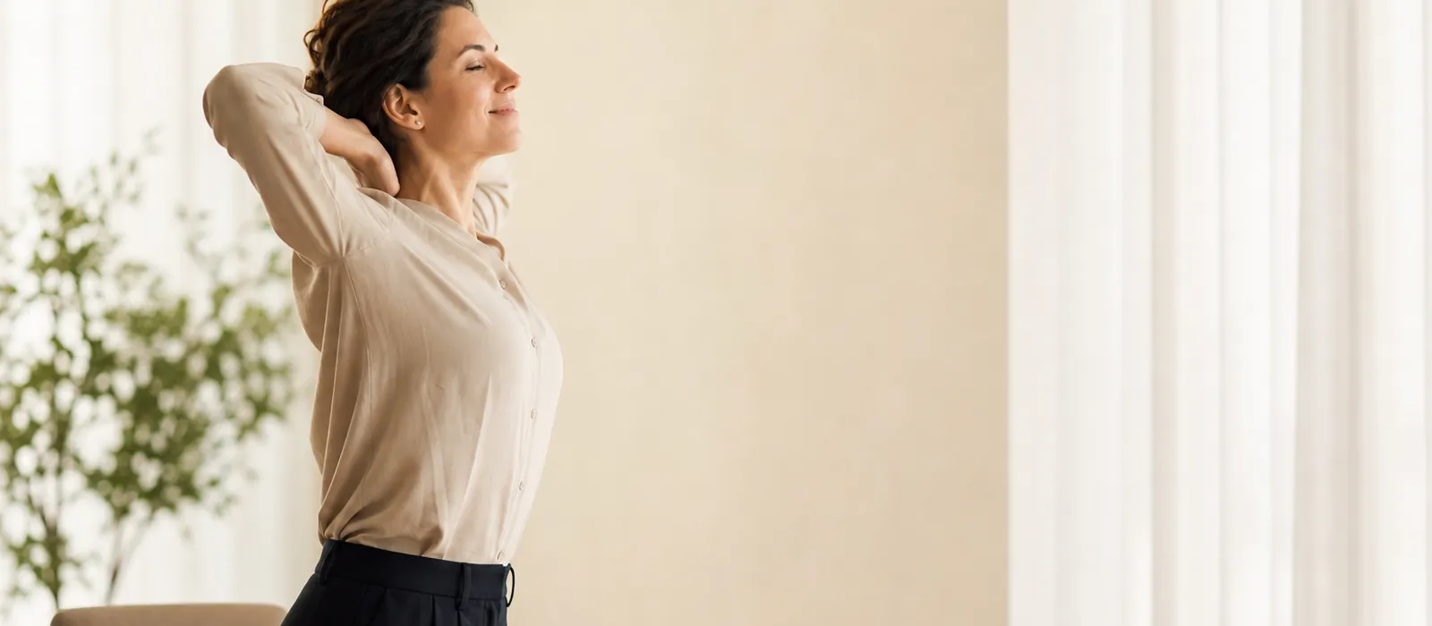 A woman standing up from her desk to stretch her upper back and neck beside a bright window