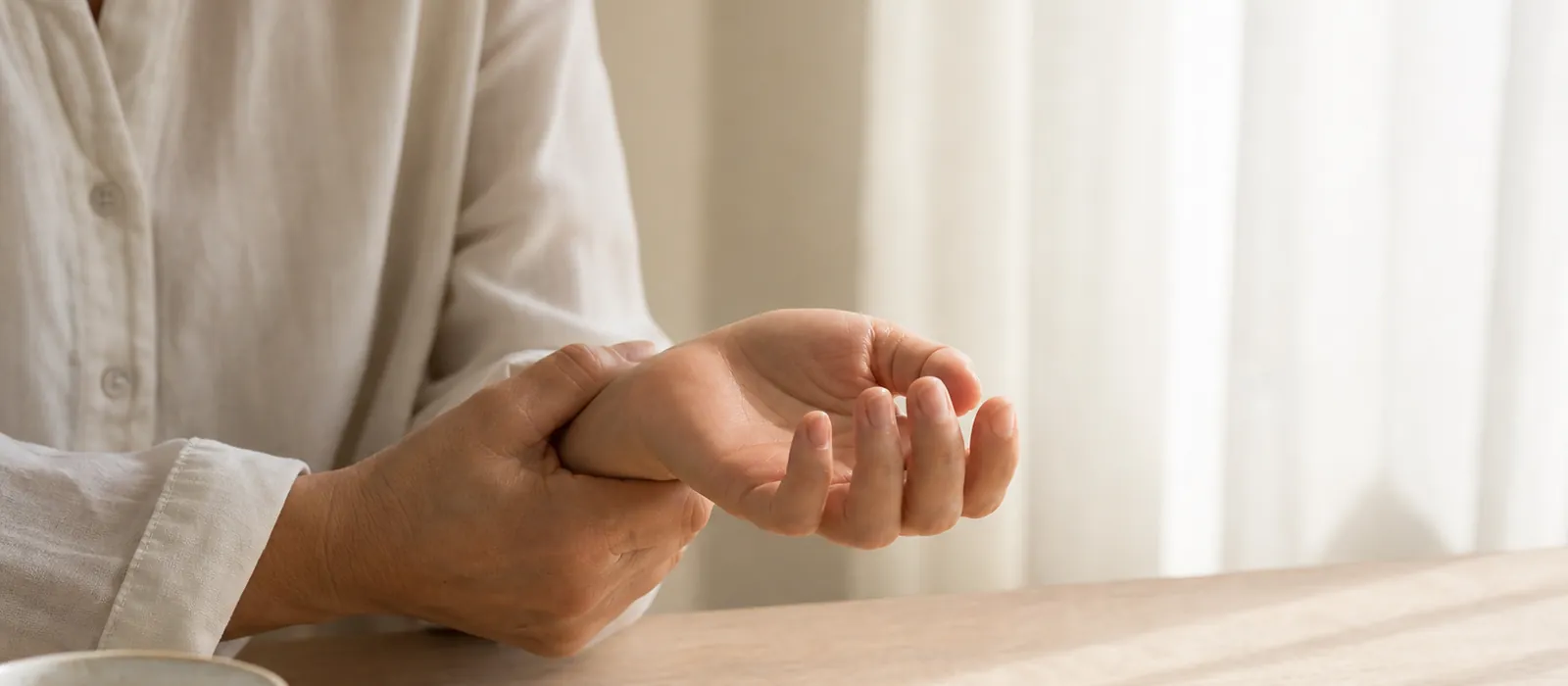 Close view of a person's hands resting on a pale oak table, gently flexing one wrist